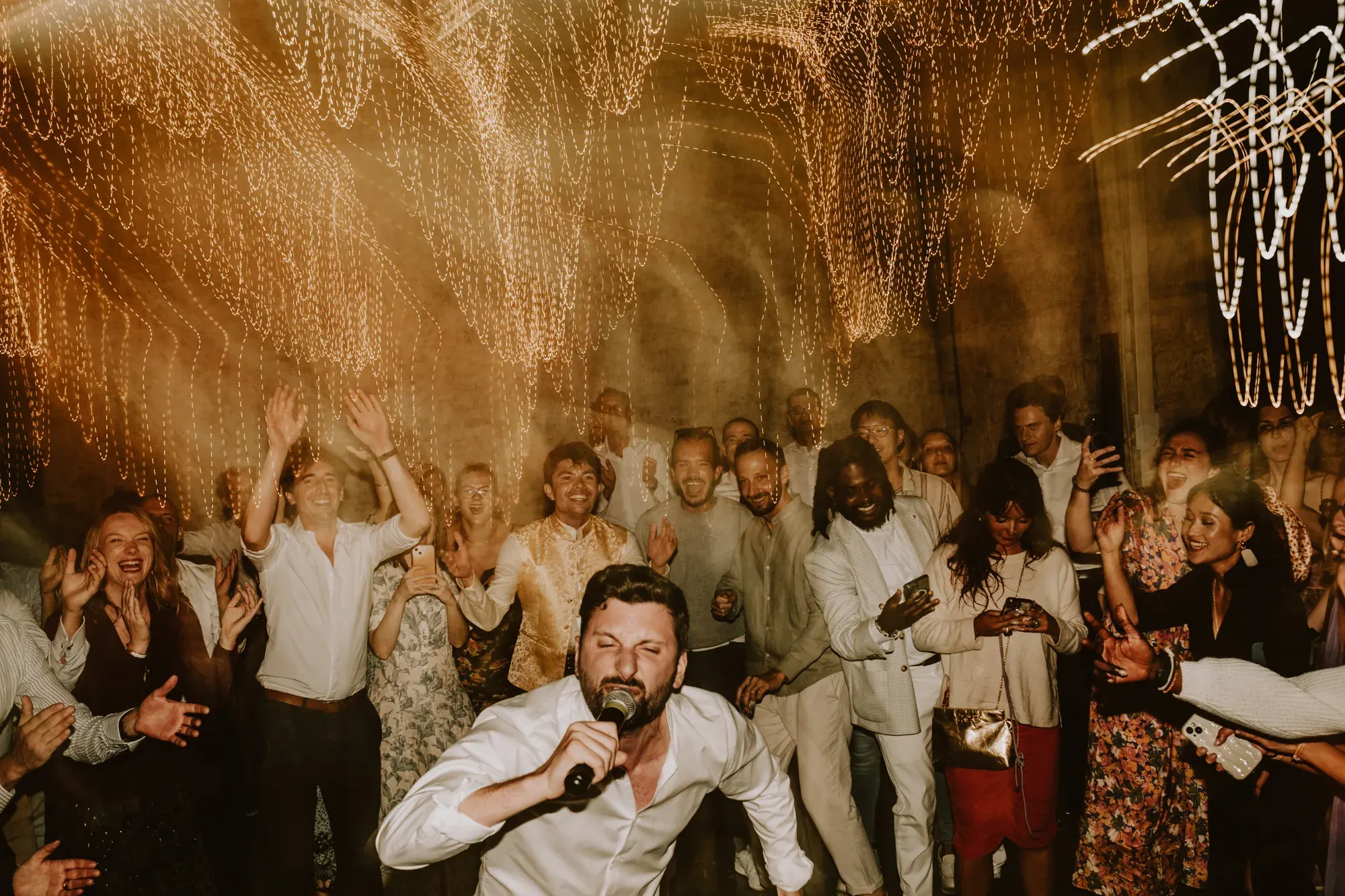 Chanteur au micro entouré des invités, guirlandes lumineuses — mariage Thiers Clermont-Ferrand, ANtO photographe